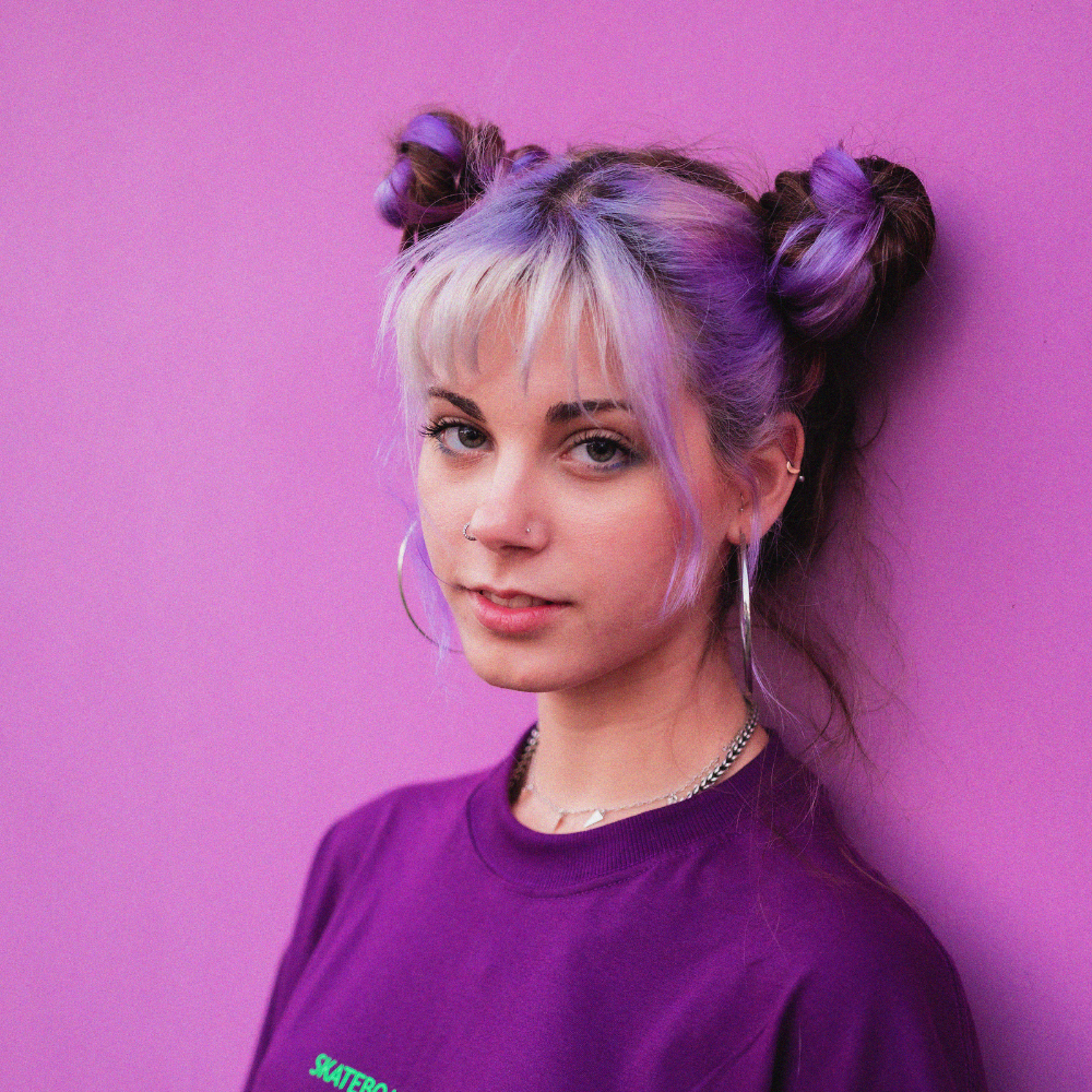 Person with purple and blonde hair styled in buns against a pink background